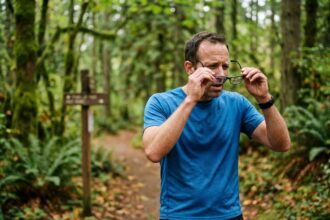 Coureur amateur sur un sentier forestier examinant ses lunettes pendant une séance d'entraînement, illustrant l'importance de la vision dans le sport amateur